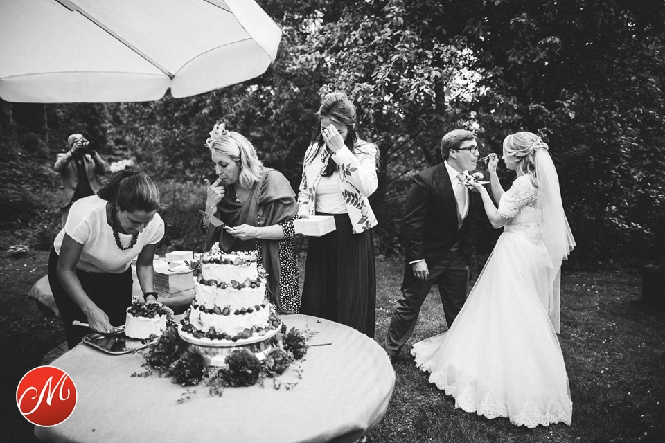 Braut füttert den Bräutigam mit Kuchen, während Gäste an der Hochzeitstorte essen