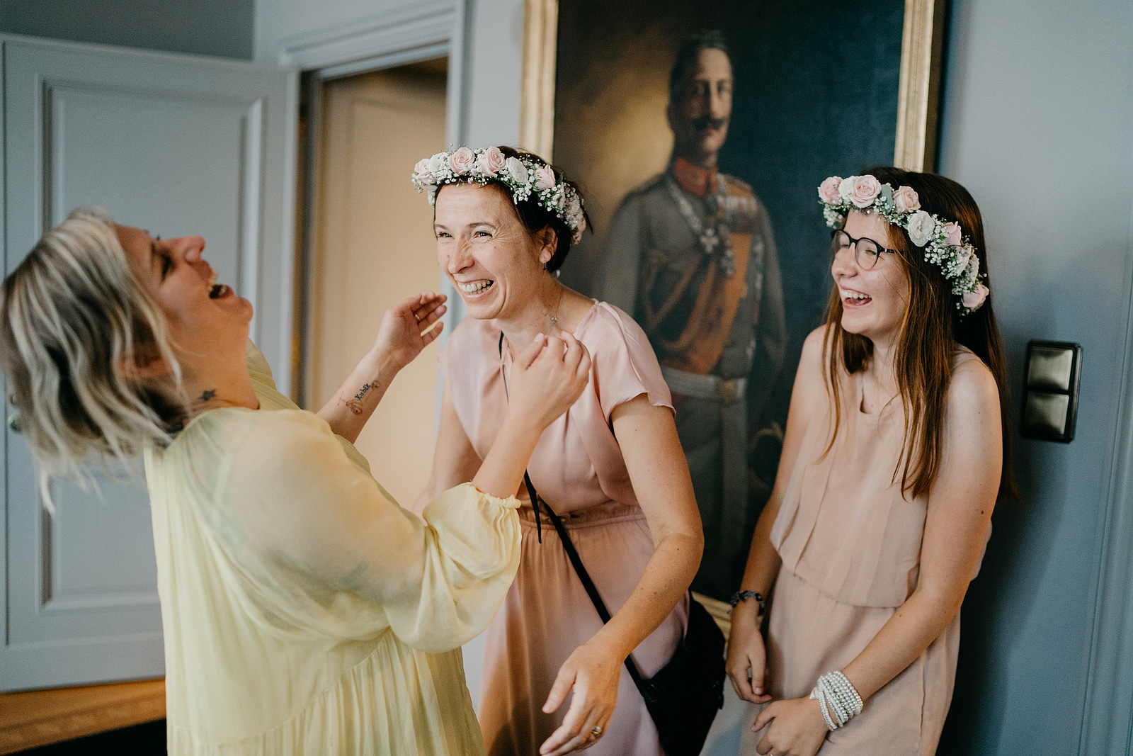 Drei Frauen lachen miteinander; zwei tragen rosa Kleider und Blumenkränze.