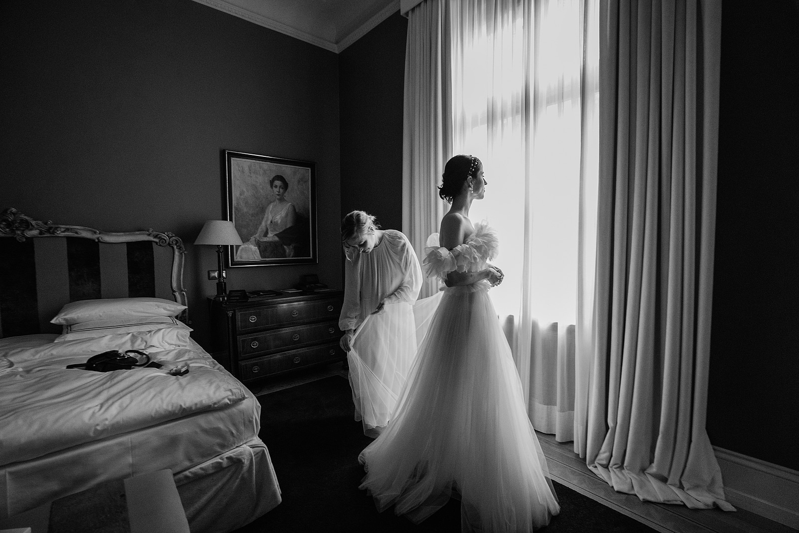 Die Braut steht im Hochzeitskleid am Fenster, während eine Frau den Rock richtet.