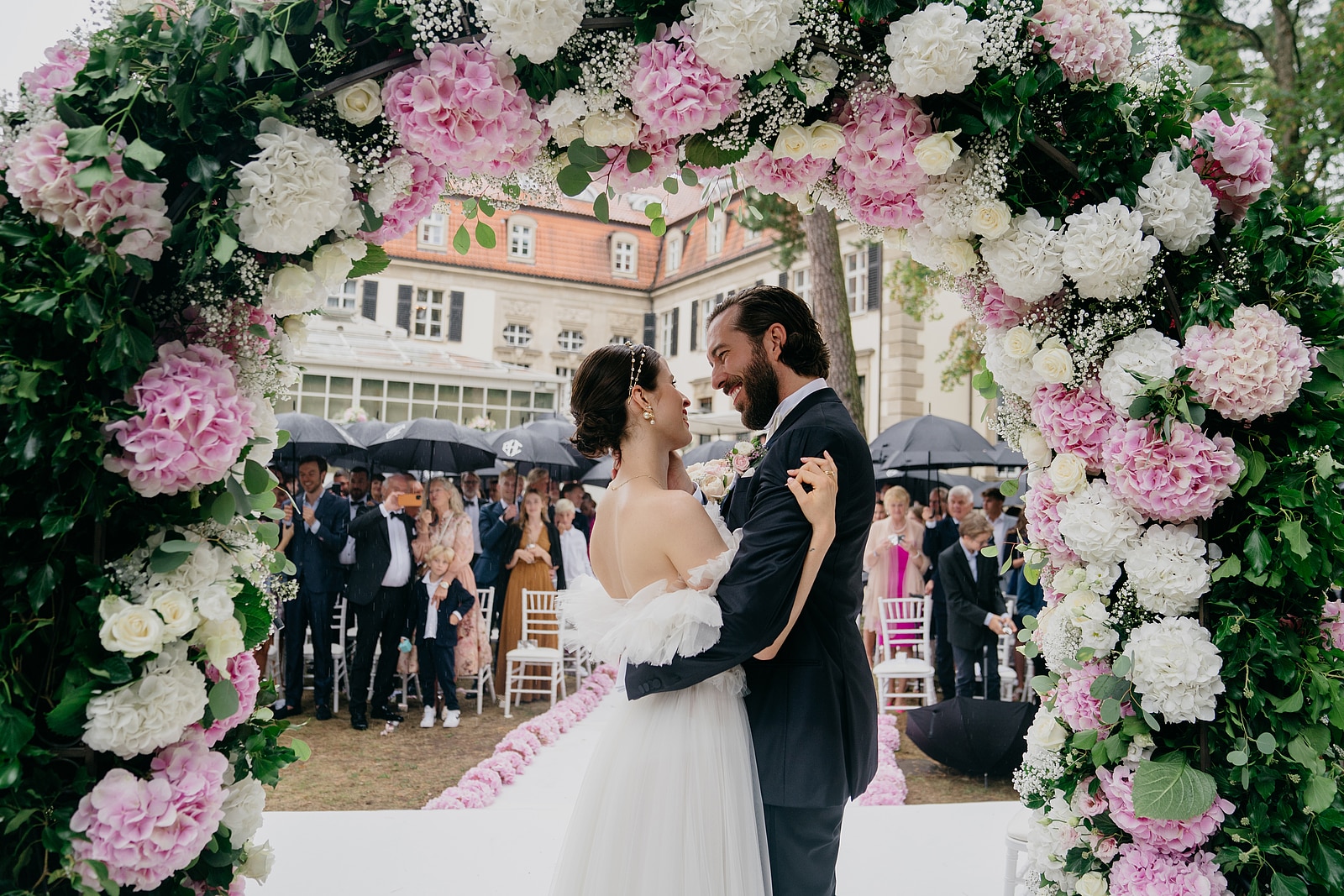 Das Brautpaar umarmt sich unter dem Blumenbogen, dahinter applaudieren die Gäste vor dem Schlosshotel Berlin.