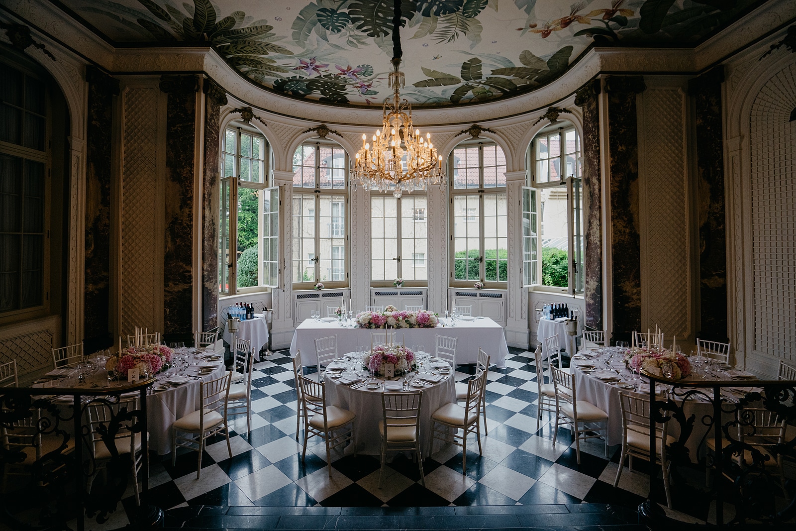 Festlich gedeckte Hochzeitstische unter einem Kronleuchter, mit rosa Blumenschmuck und schwarz-weißem Fliesenboden.