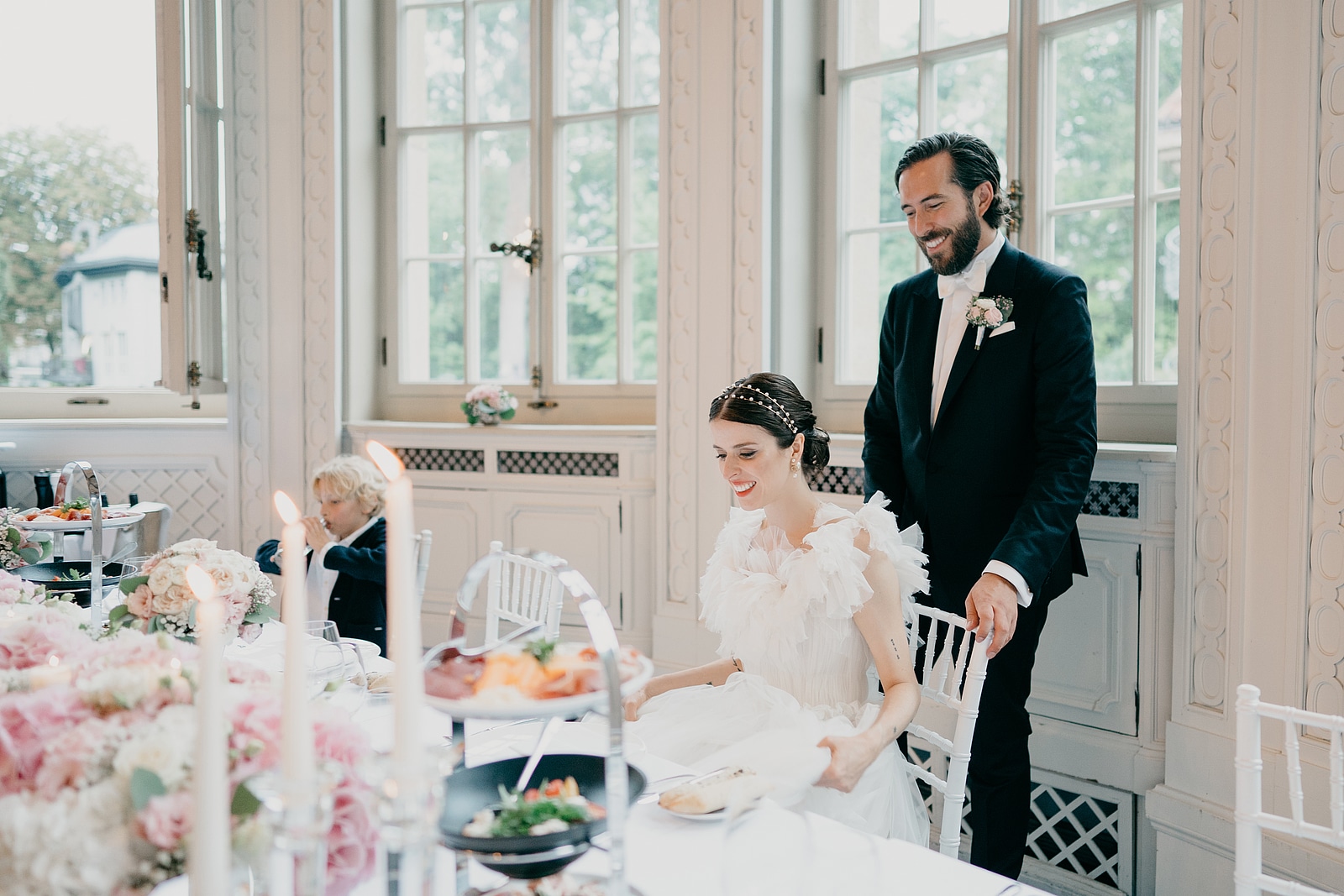 Die Braut nimmt am gedeckten Hochzeitstisch Platz, der Bräutigam steht hinter ihrem Stuhl.