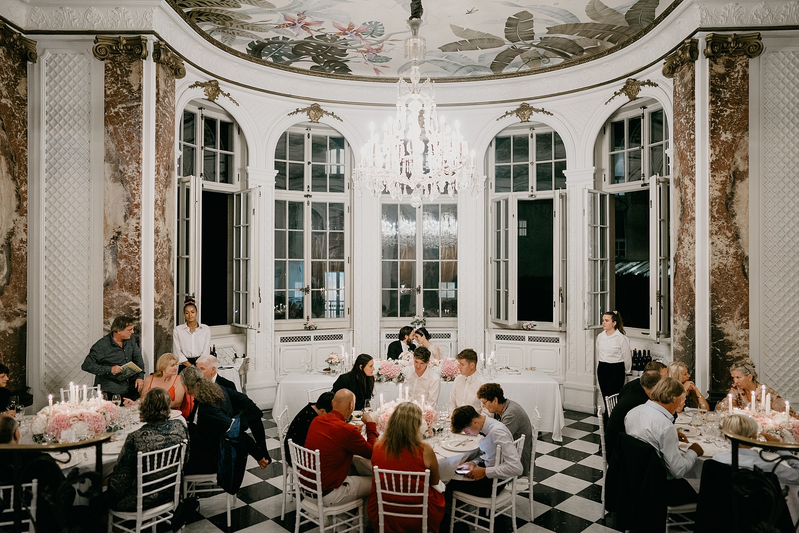 Die Hochzeitsgesellschaft sitzt zum Abendessen an runden Tischen unter dem großen Kronleuchter.